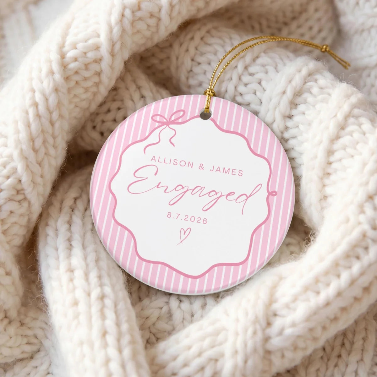 Pink pinstripe round ceramic Engaged ornament with personalized names and date resting in a cream knit blanket