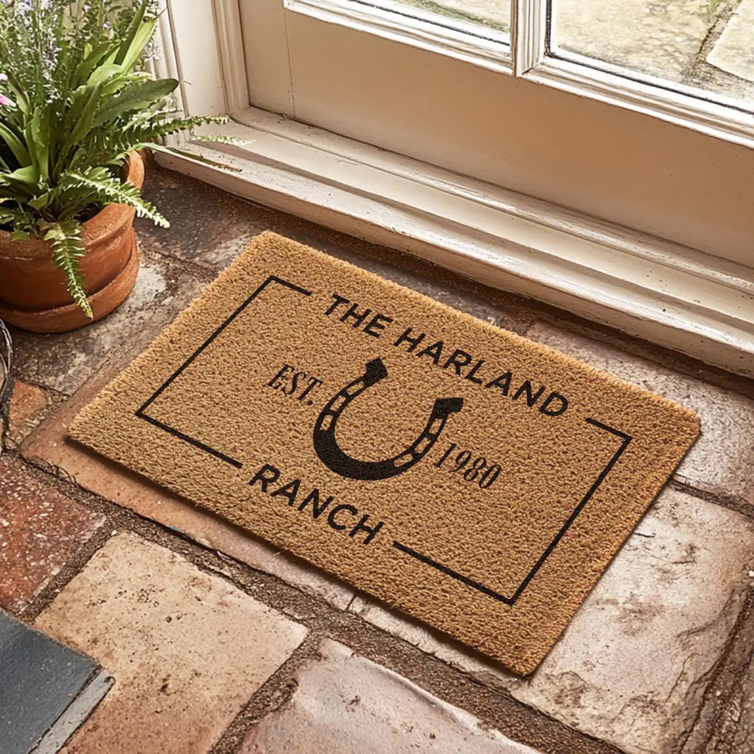 Coir doormat with a horseshoe ranch design on a stone floor beside a window and potted fern