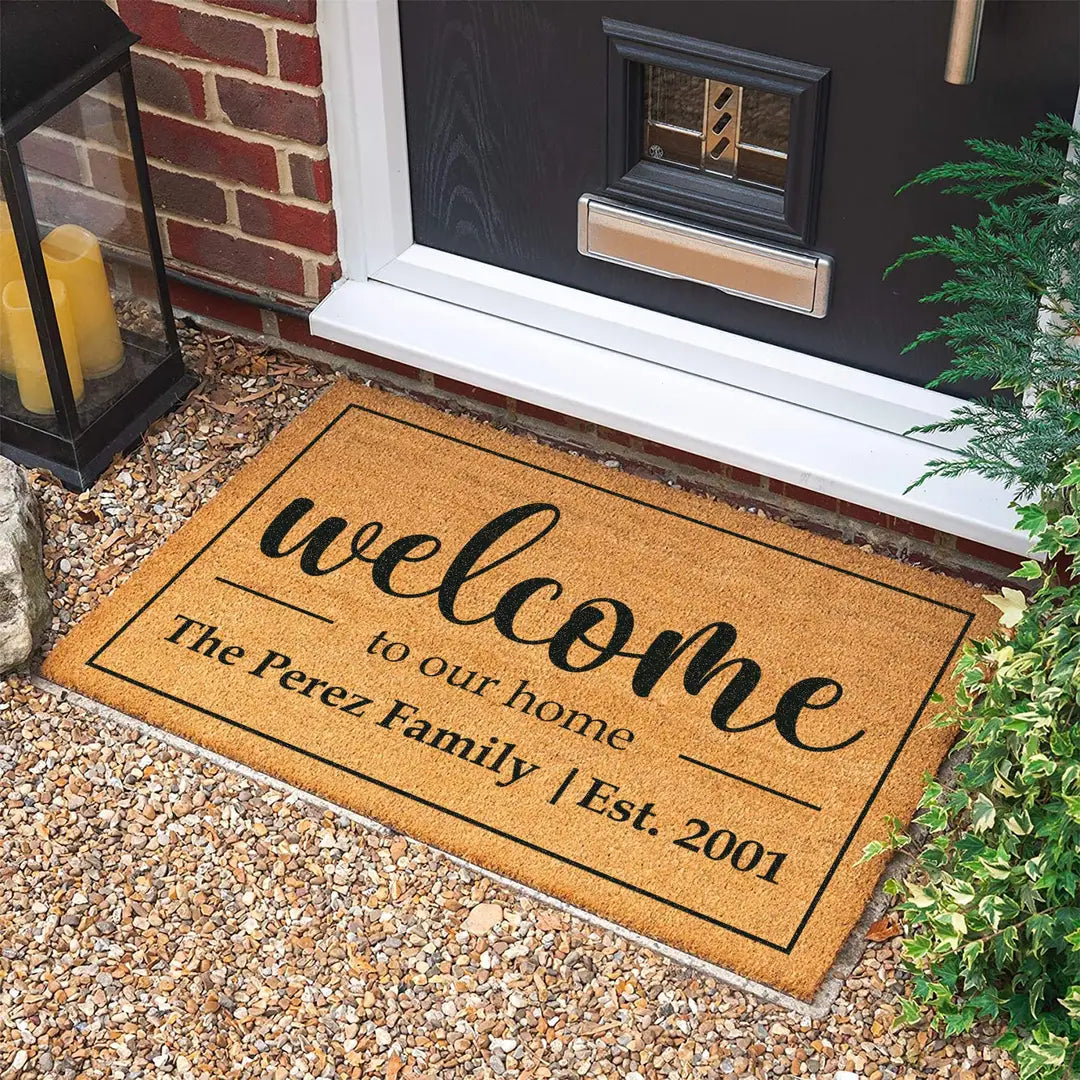 Rectangular coir doormat with a bordered welcome to our home design and family name at a dark front door on gravel