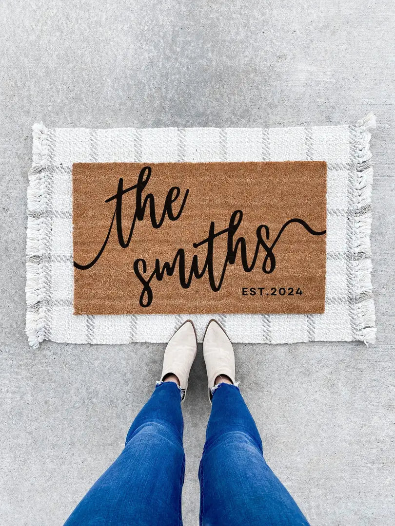 Coir doormat with a script family name and established year layered on a white fringed rug with a person in jeans and boots standing on concrete