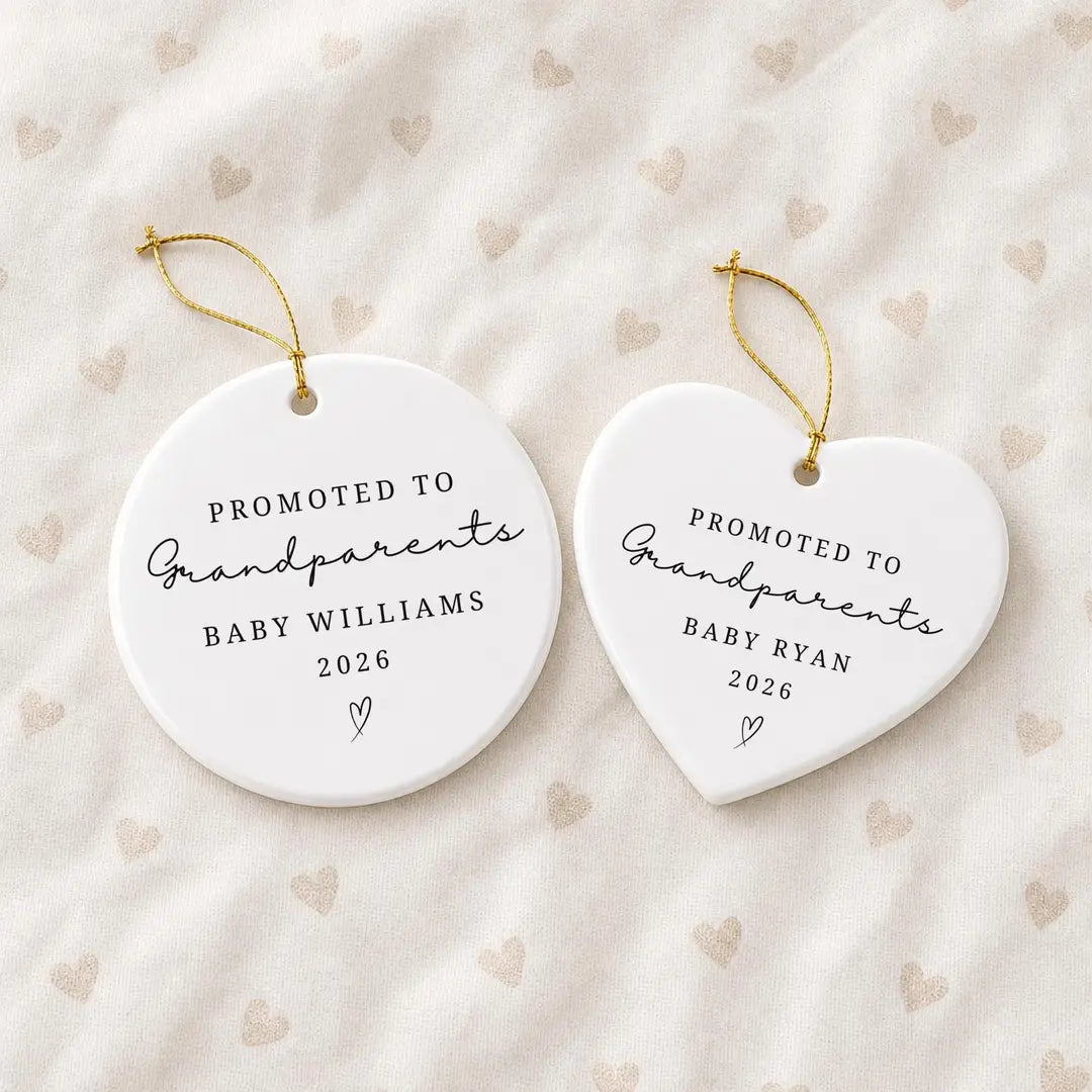 Round and heart-shaped white ceramic Promoted to Grandparents ornaments with minimal script lettering, a baby name and year, and gold hanging strings on a heart-print fabric