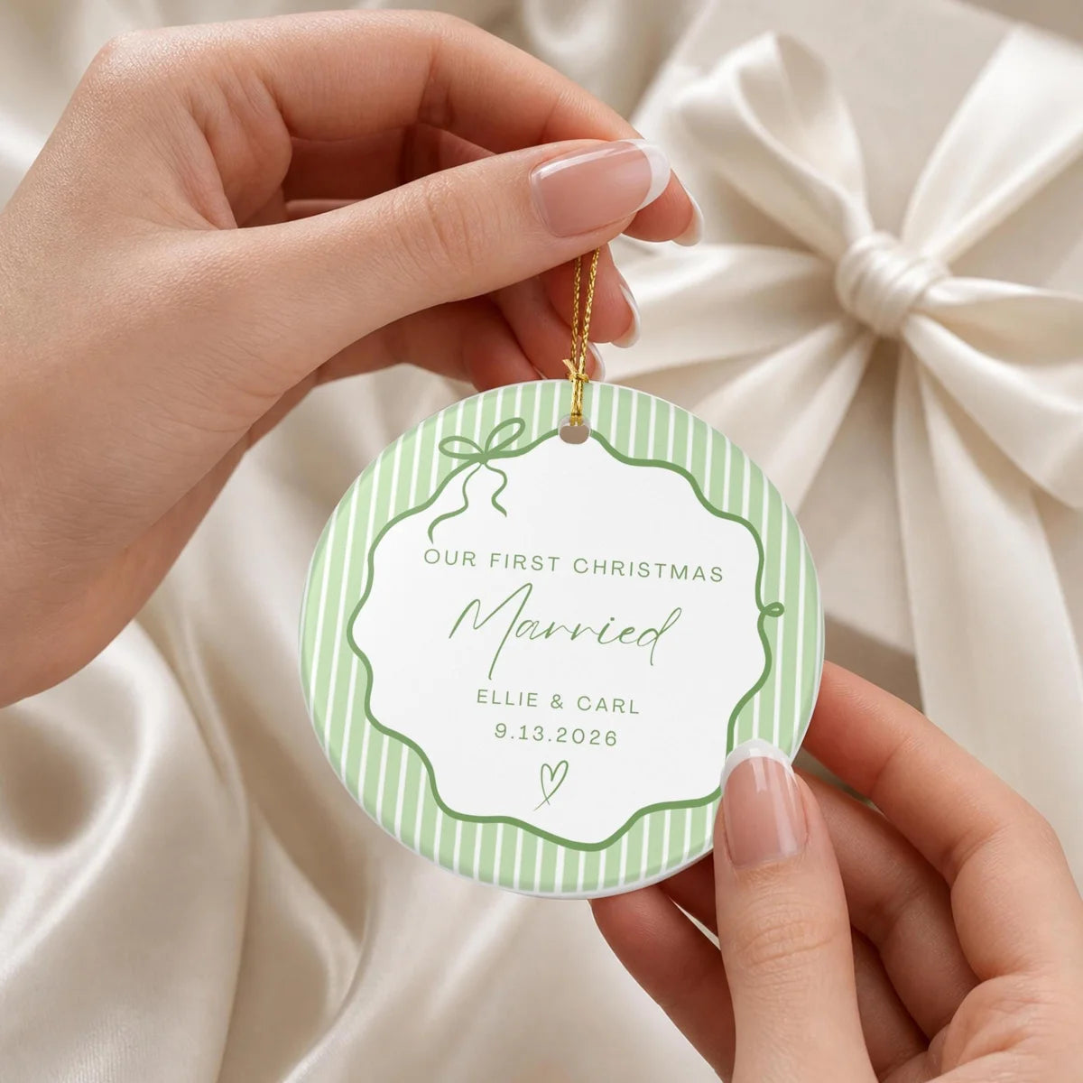 Hands holding a green pinstripe round ceramic First Christmas Married ornament by its gold string in front of a cream gift bow