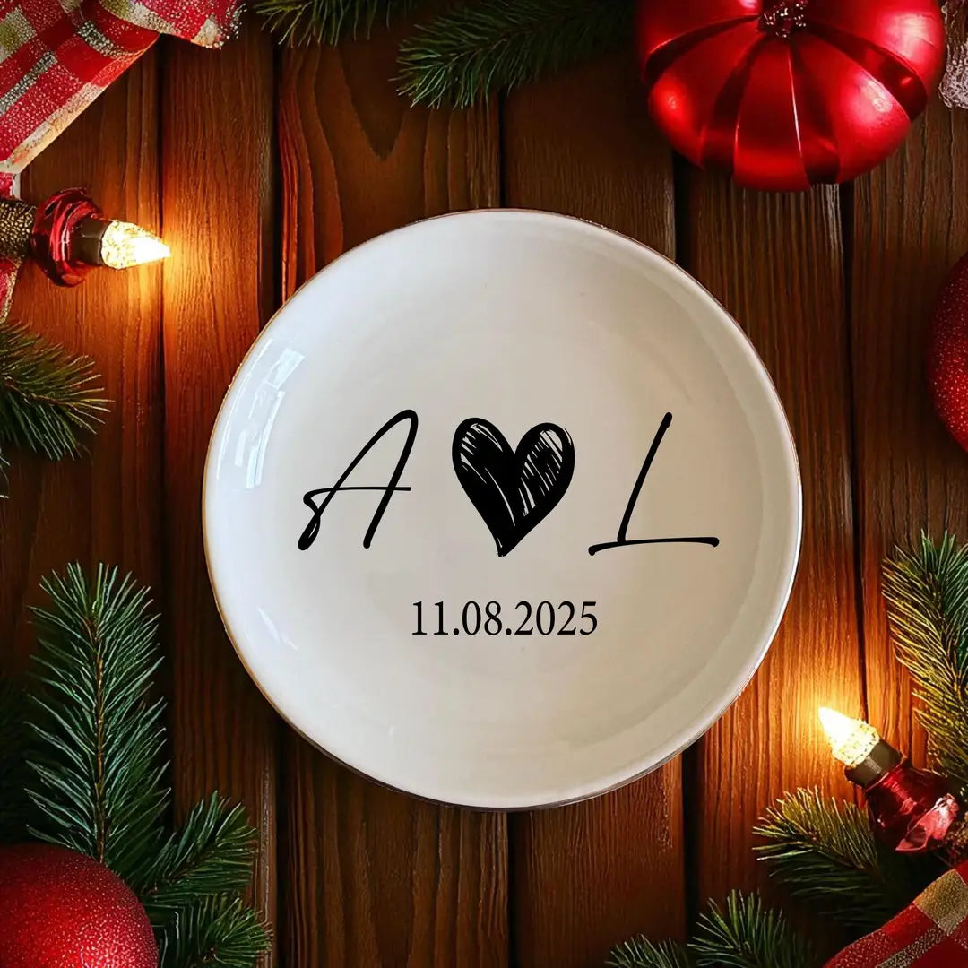 Round ceramic ring dish with black script initials and a date, styled on wood with holiday greenery and red ornaments