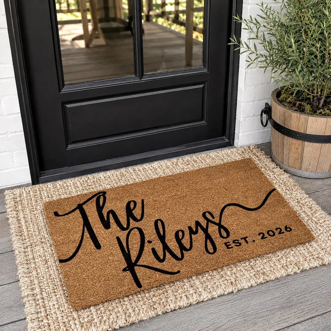 Coir doormat with a script family name and established year layered on a jute rug at a black door with a potted olive tree