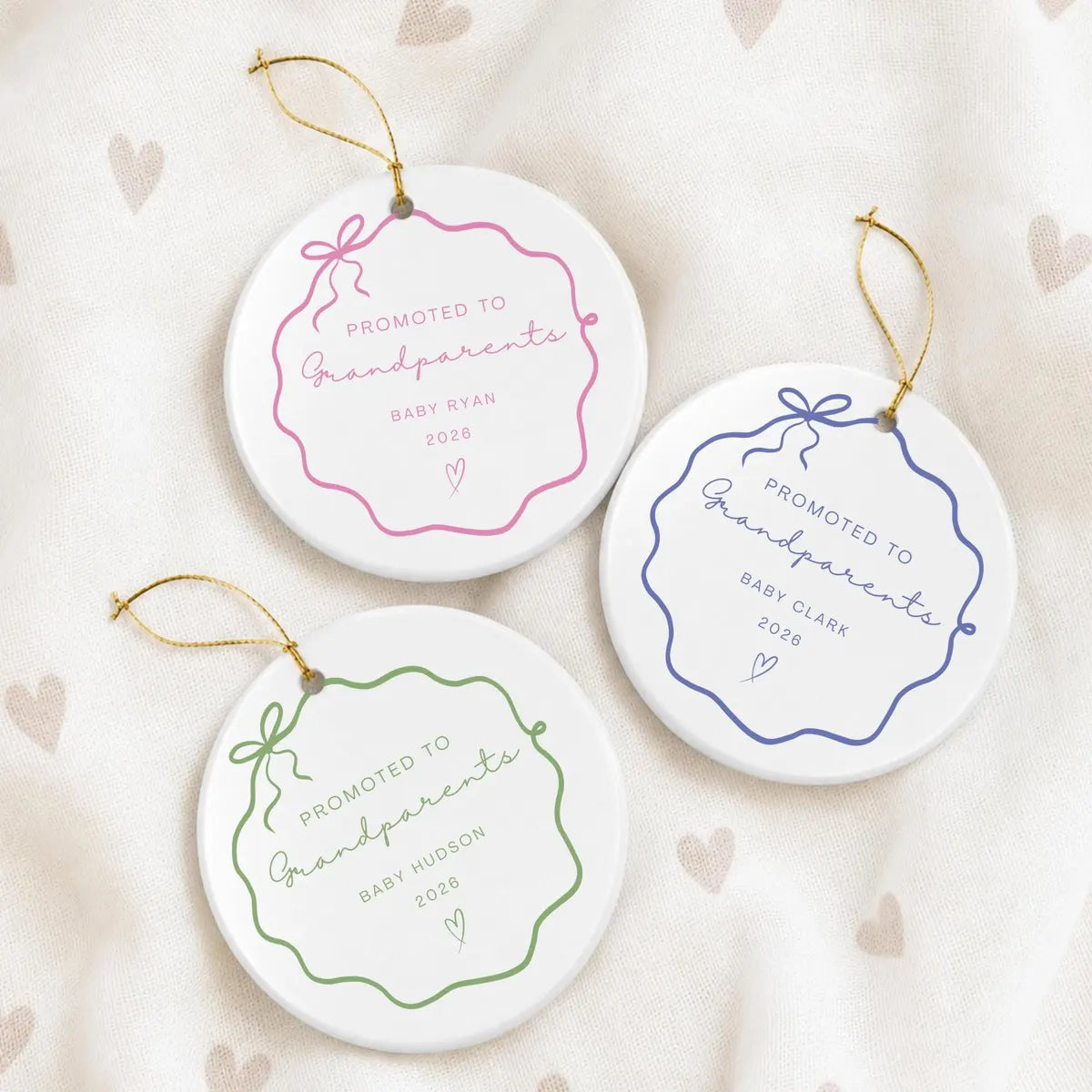 Pink, blue, and green wavy bow frame round ceramic Promoted to Grandparents ornaments on a cream heart-print fabric