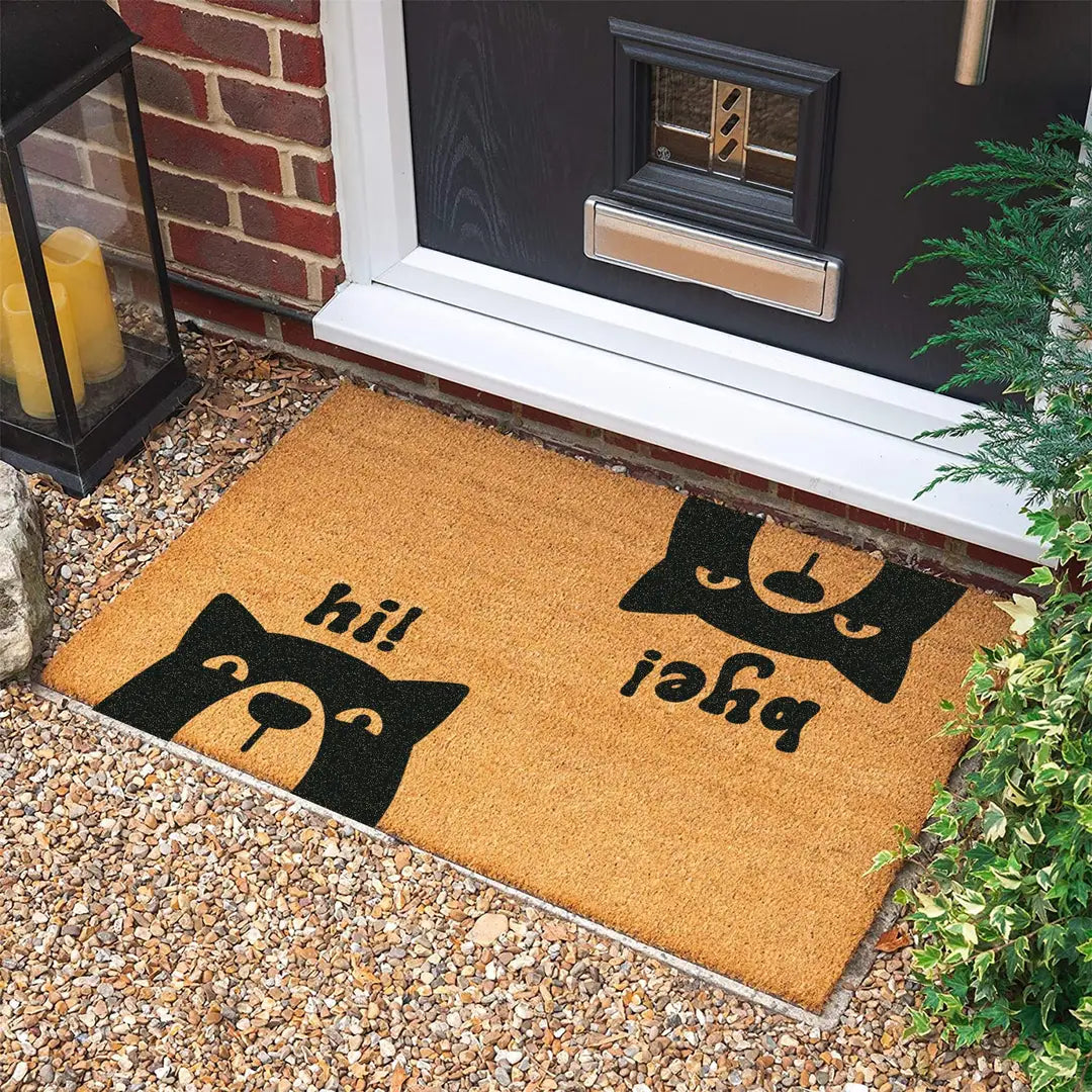 Rectangular coir doormat with two dog faces and hi and bye lettering at a dark front door on gravel