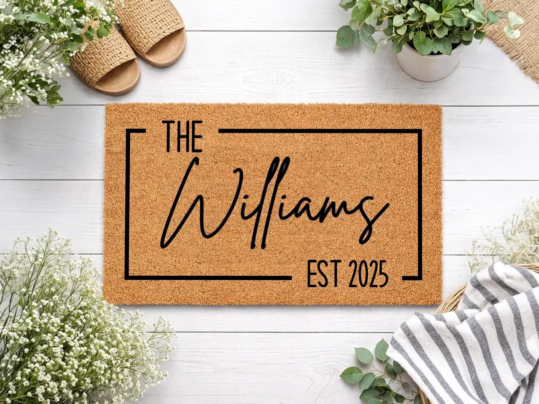 Coir doormat with a framed family name and established year on white boards with straw sandals, baby's breath, and a striped towel
