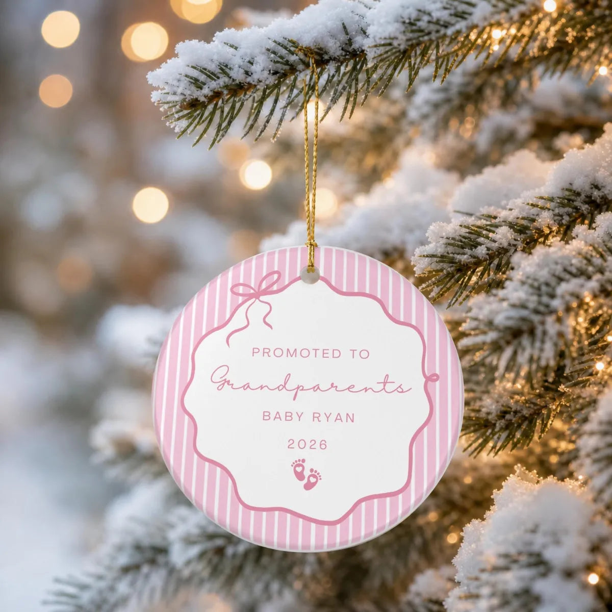 Pink pinstripe round ceramic Promoted to Grandparents ornament hanging from a snow-covered evergreen branch on a gold string