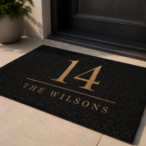 Black faux coir doormat with a gold-tone house number and family name on a stone step at a dark door