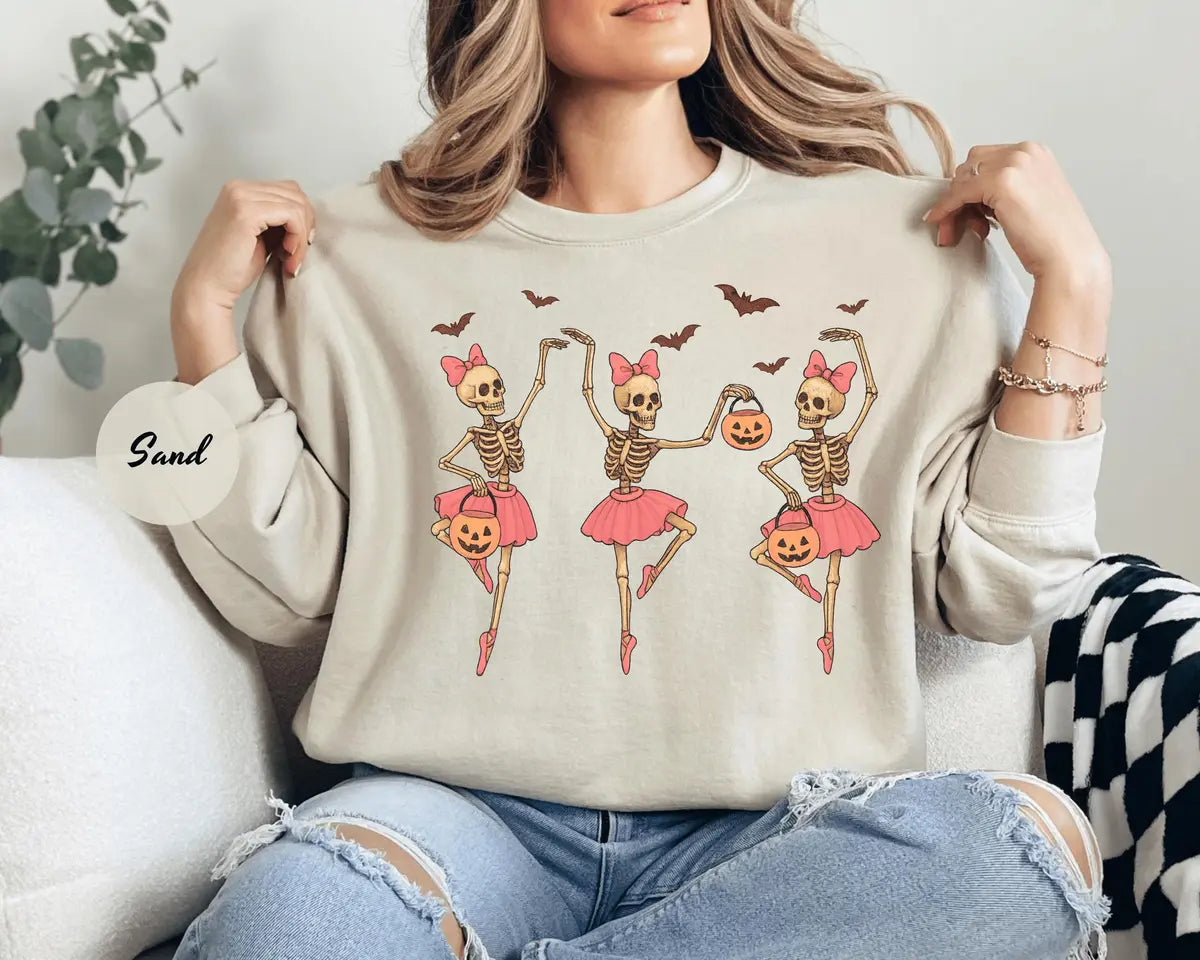 Sand crewneck sweatshirt with printed three ballerina skeletons in pink tutus with pumpkins and bats, worn by a model.