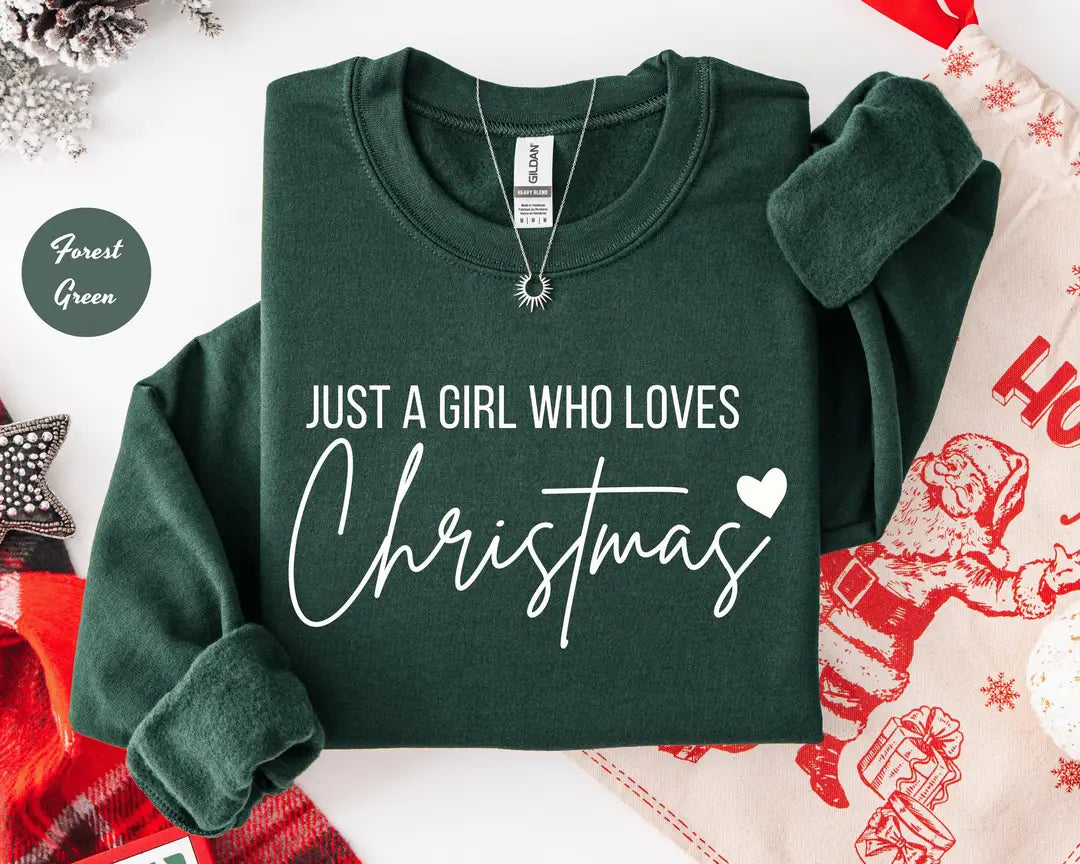 Forest Green crewneck sweatshirt with script lettering that reads just a girl who loves Christmas with a small heart, flat lay view.