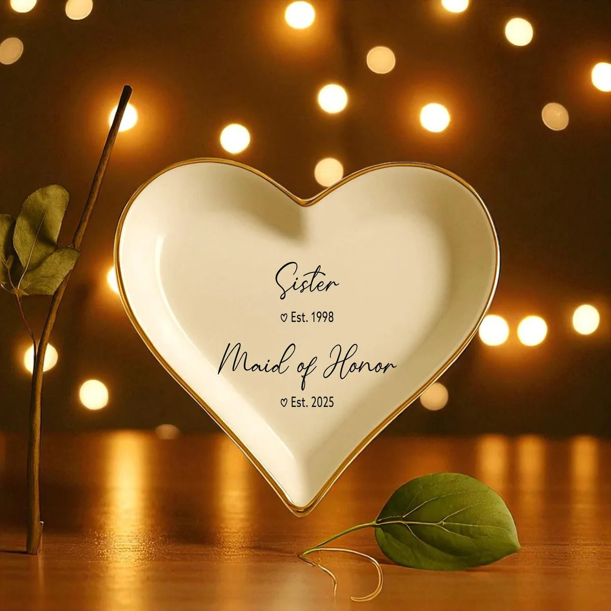 Heart-shaped ceramic maid of honor ring dish with gold rim and Sister and Maid of Honor years, styled with a leaf stem and warm glowing lights