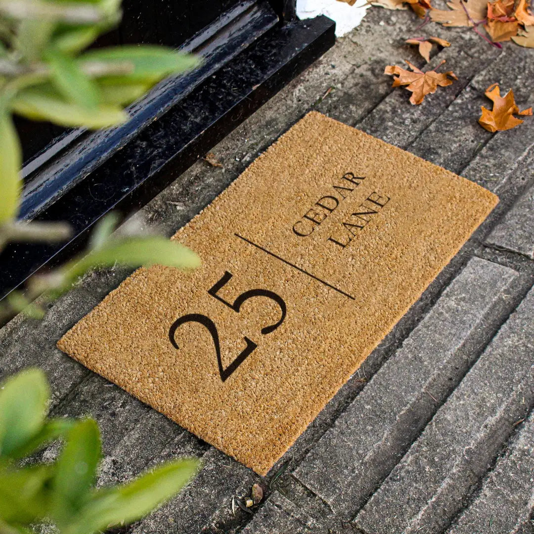Coir doormat with a house number and street name on dark stone steps with autumn leaves and green foliage