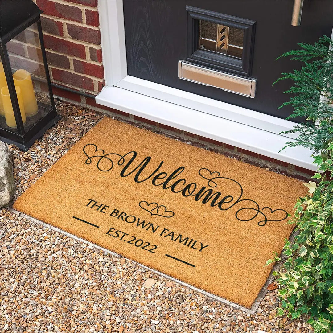 Rectangular coir doormat with a heart script Welcome, family name, and established year on gravel in front of a black door