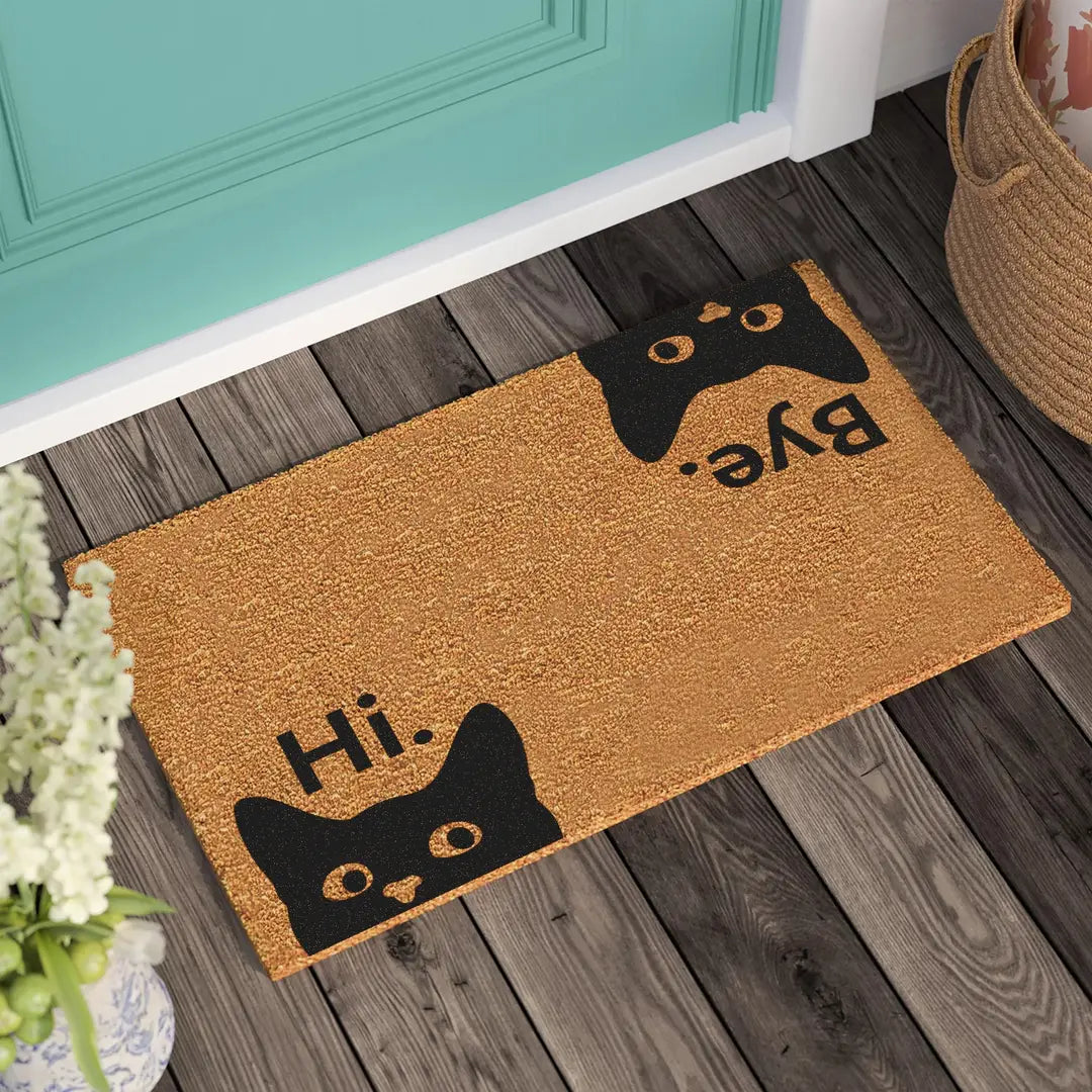 Coir doormat with Hi and Bye lettering and two peeking black cats on a wooden porch beside a teal door and woven basket