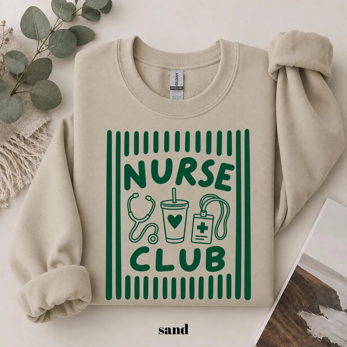 Sand crewneck sweatshirt with printed a retro striped frame with a stethoscope, coffee cup and badge above the words nurse club, flat lay view.