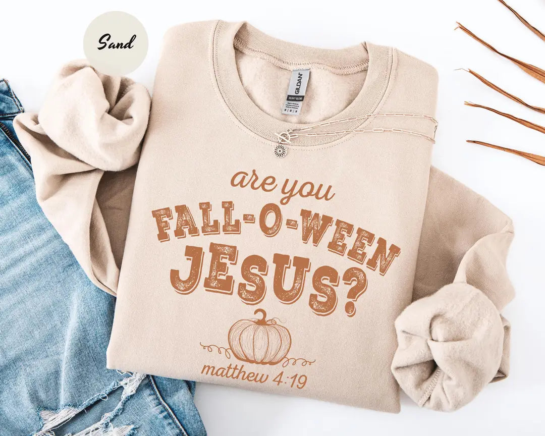 Sand crewneck sweatshirt with printed a pumpkin with the words are you fall-o-ween jesus and matthew 4:19 in a western style font, flat lay view.