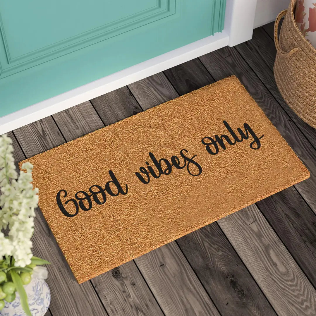 Coir doormat with Good Vibes Only cursive lettering on a wooden porch beside a teal door and woven basket