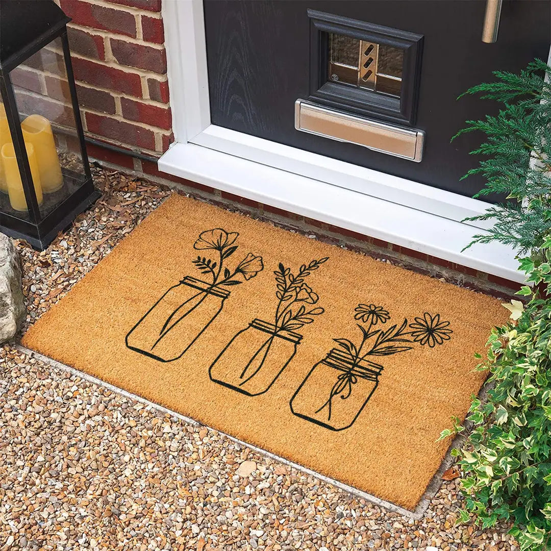 Rectangular coir doormat with three line art wildflower bouquets in jars at a dark front door on gravel