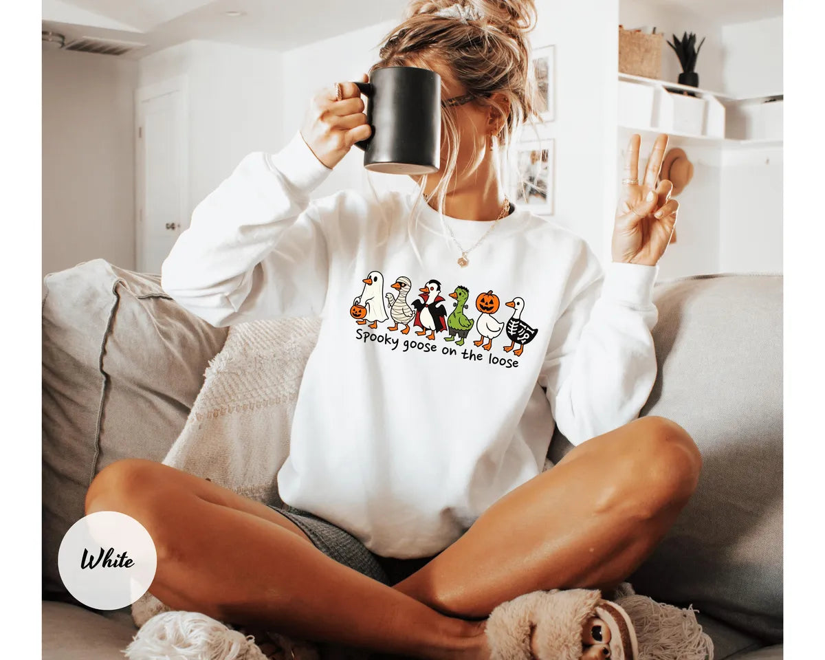 White crewneck sweatshirt with costumed geese dressed as a ghost, vampire, pumpkin and skeleton above the words spooky goose on the loose, worn by a model.