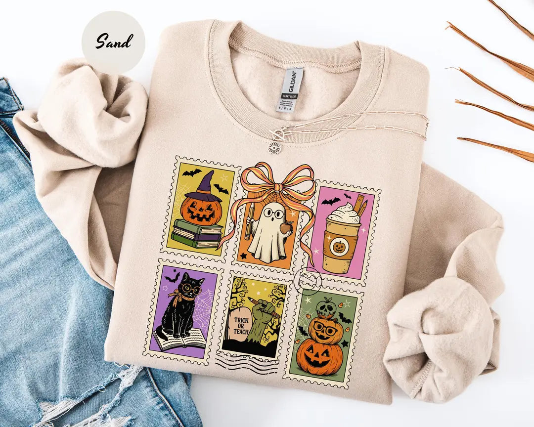 Sand crewneck sweatshirt with six vintage postage-style stamps with a ghost, black cat, pumpkins and a coffee cup, flat lay view.
