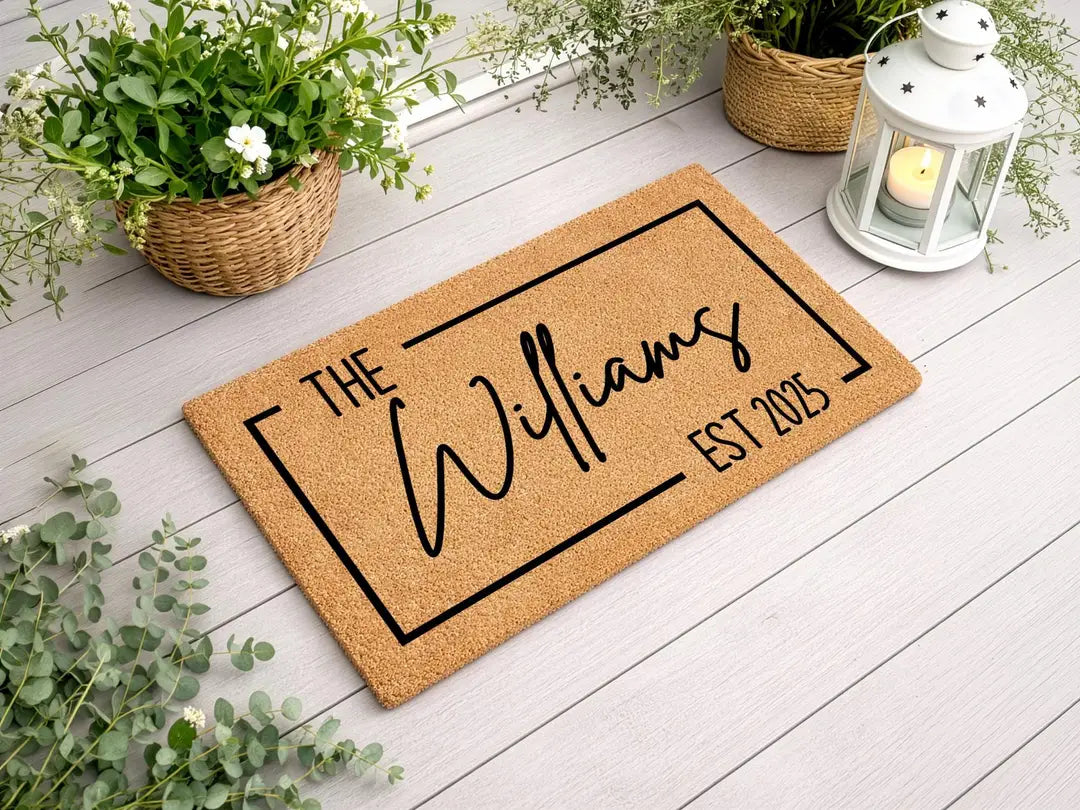 Coir doormat with a framed family name and established year on a white wood deck with a lantern and flower baskets