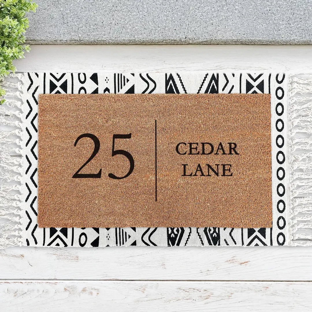 Coir doormat with a house number and street name layered on a black and white geometric fringed rug on white boards