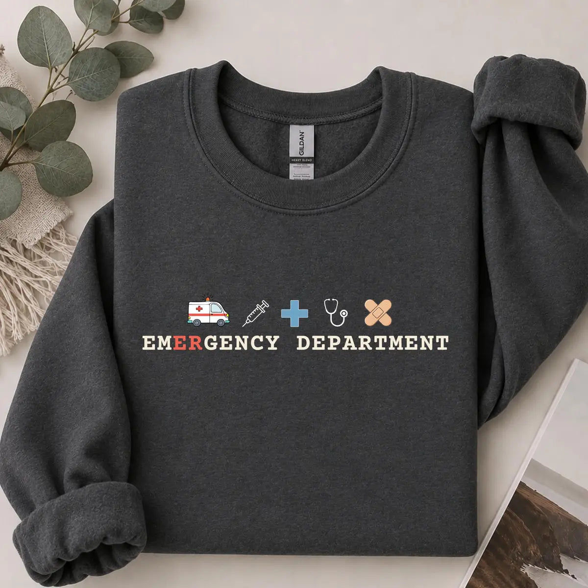 Dark Heather crewneck sweatshirt with printed an ambulance, syringe, cross, stethoscope and bandage above the words emergency department, flat lay view.