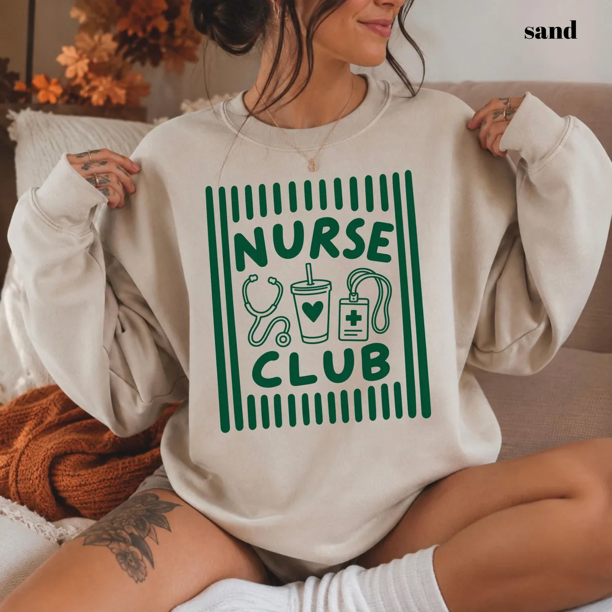 Sand crewneck sweatshirt with printed a retro striped frame with a stethoscope, coffee cup and badge above the words nurse club, worn by a model.