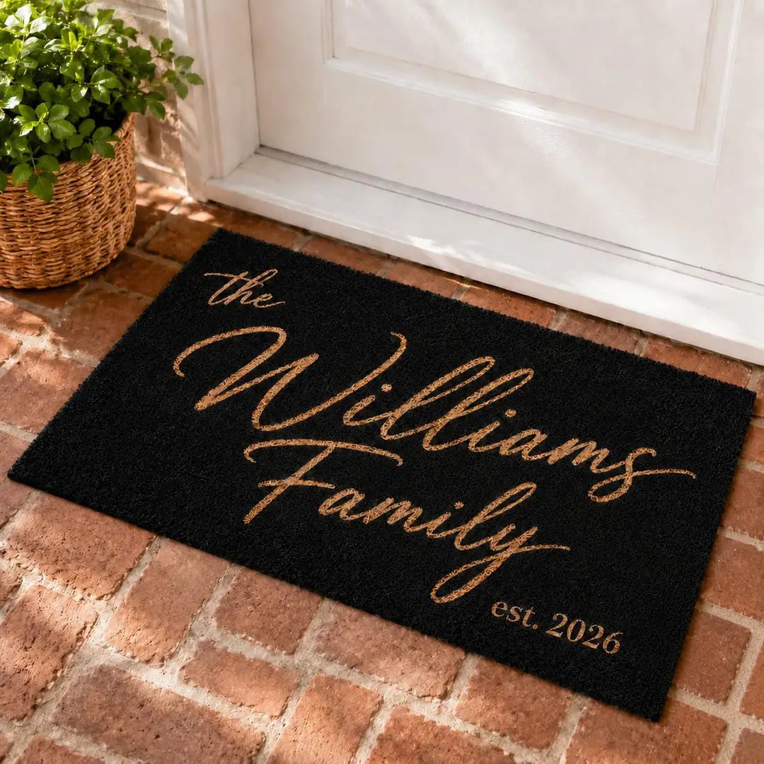 Rectangular black faux coir doormat with a gold-tone script family name and established year at a white door on brick