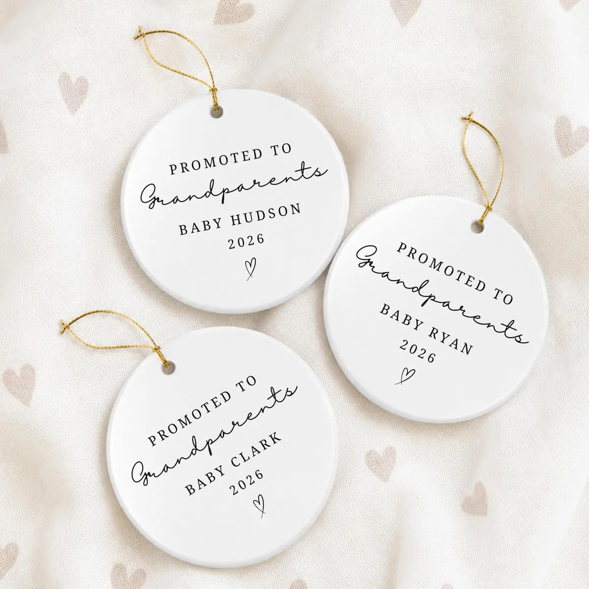 Three round white ceramic Promoted to Grandparents ornaments with different personalized baby names and the year on a heart-print fabric