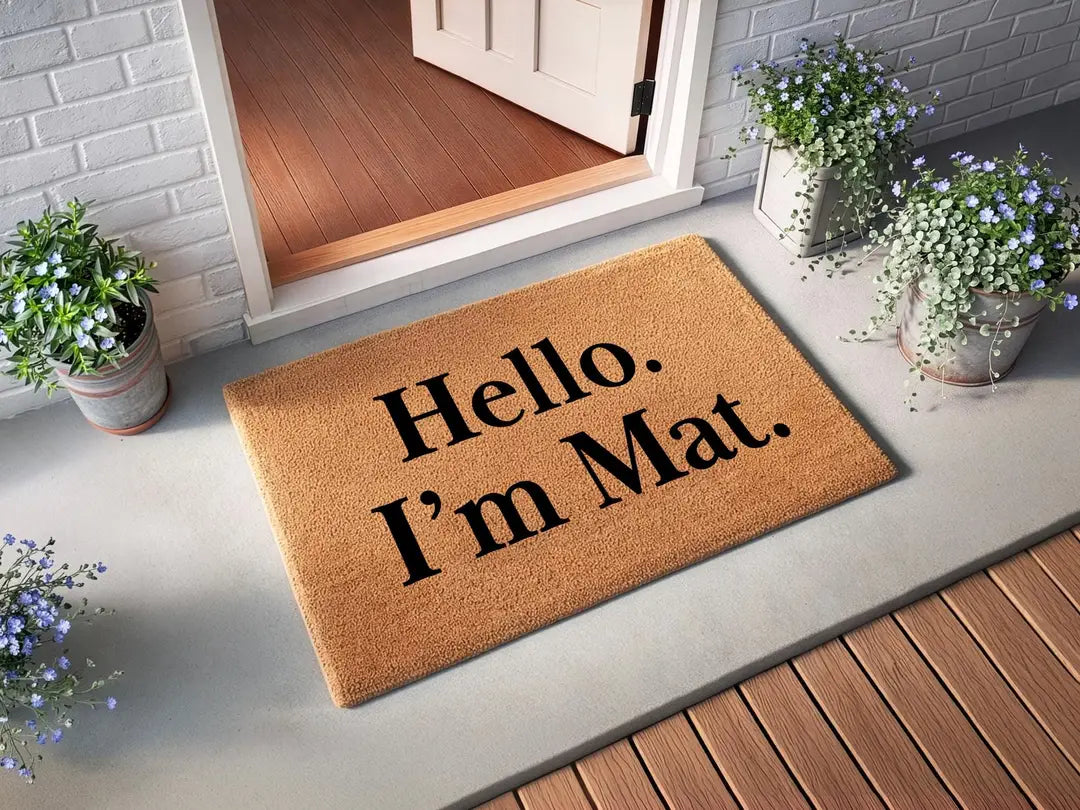 Rectangular coir doormat with a Hello I'm Mat message at an open white door with white brick siding and potted blue flowers