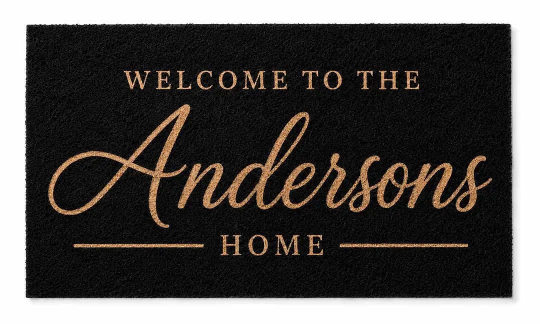 Flat view of a black faux coir doormat with Welcome to the, a gold-tone script surname, and a Home line