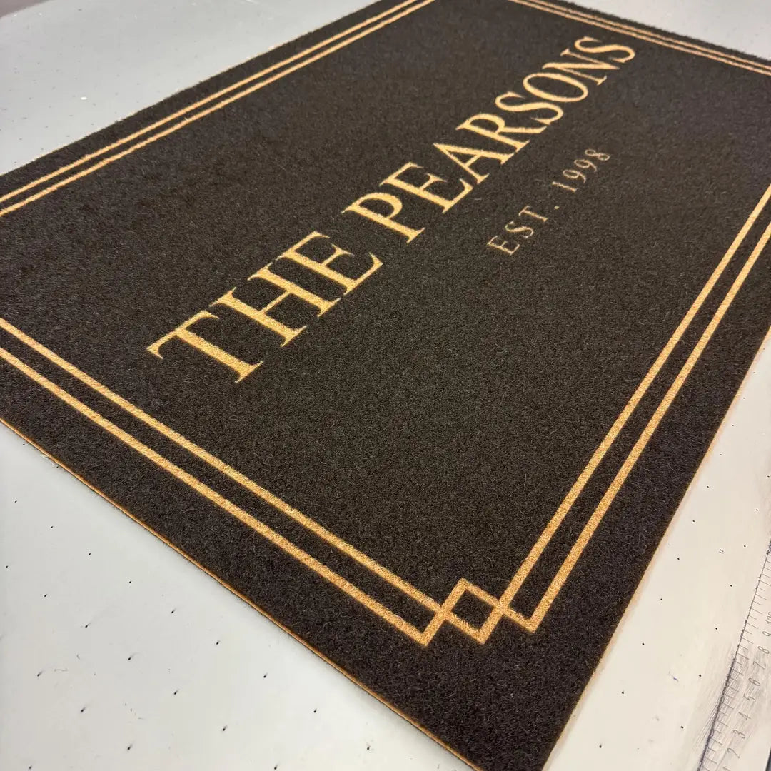 Angled closeup of a dark faux coir doormat with a double gold-tone border, family name, and established year