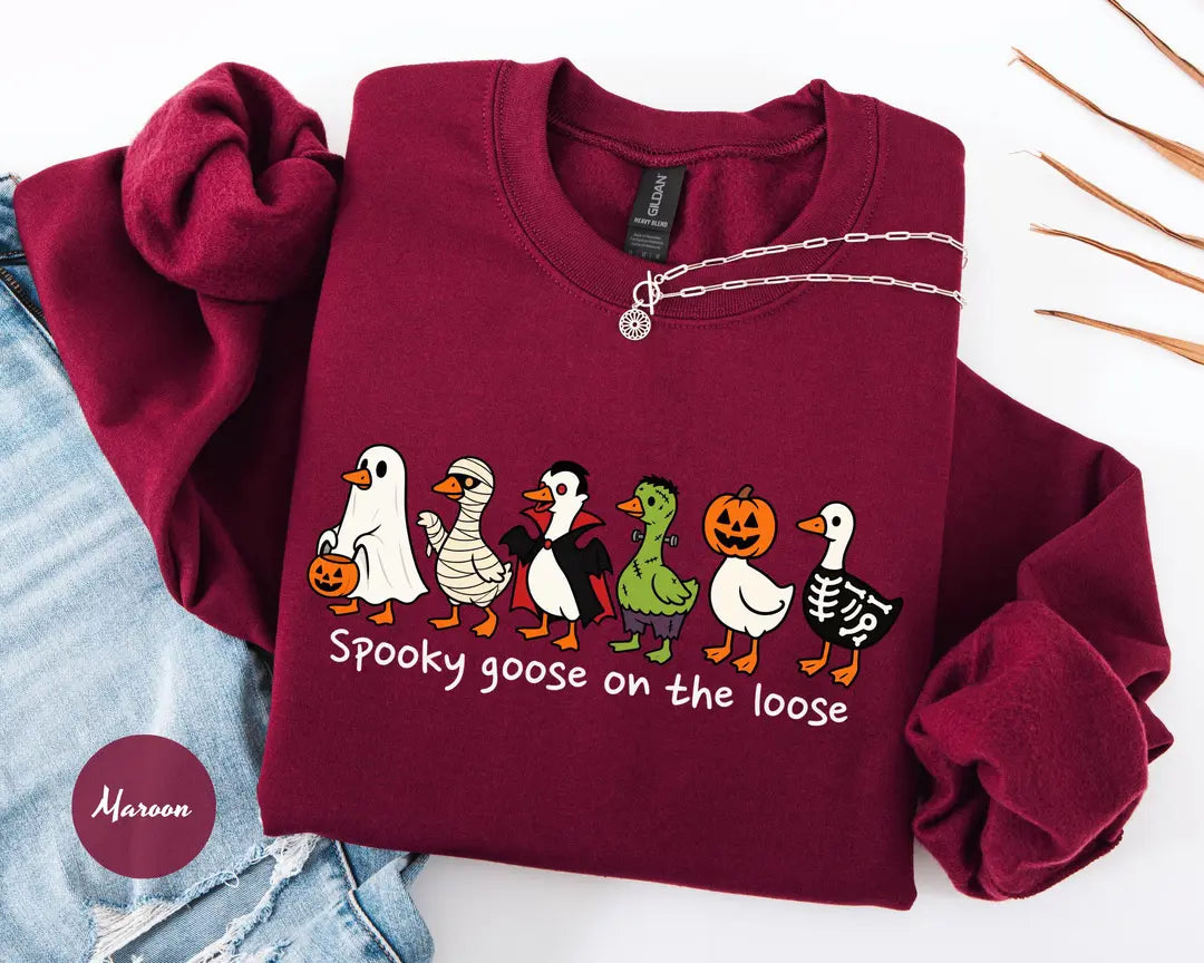 Maroon crewneck sweatshirt with costumed geese dressed as a ghost, vampire, pumpkin and skeleton above the words spooky goose on the loose, flat lay view.