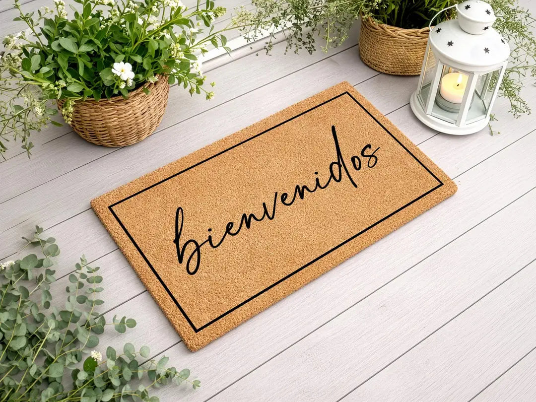 Coir doormat with a framed bienvenidos script message on a white wood deck with a lantern and flower baskets