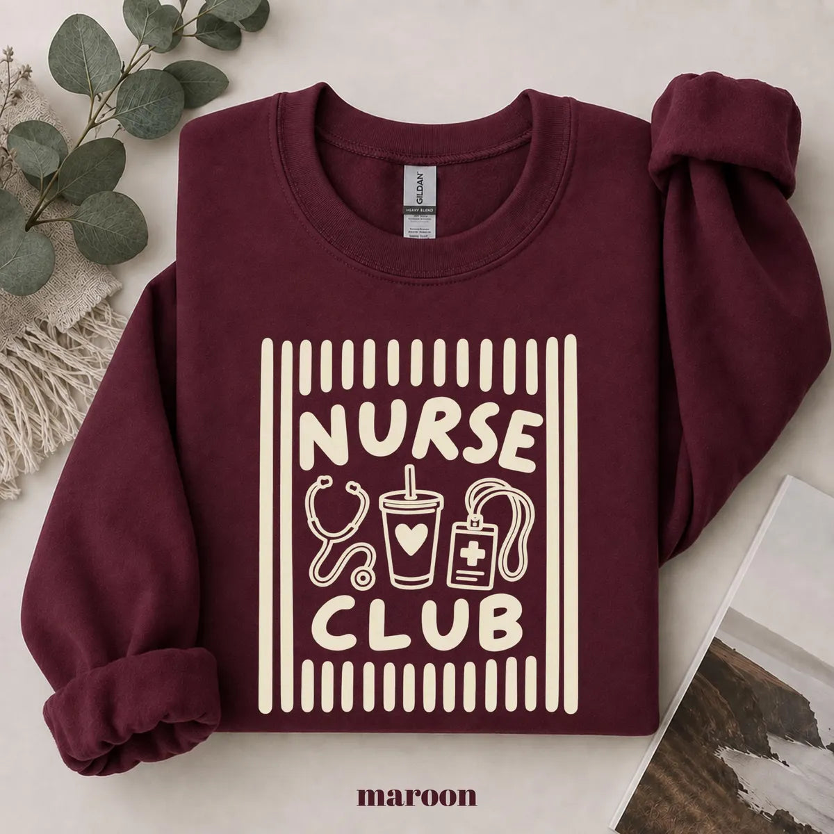 Maroon crewneck sweatshirt with printed a retro striped frame with a stethoscope, coffee cup and badge above the words nurse club, flat lay view.