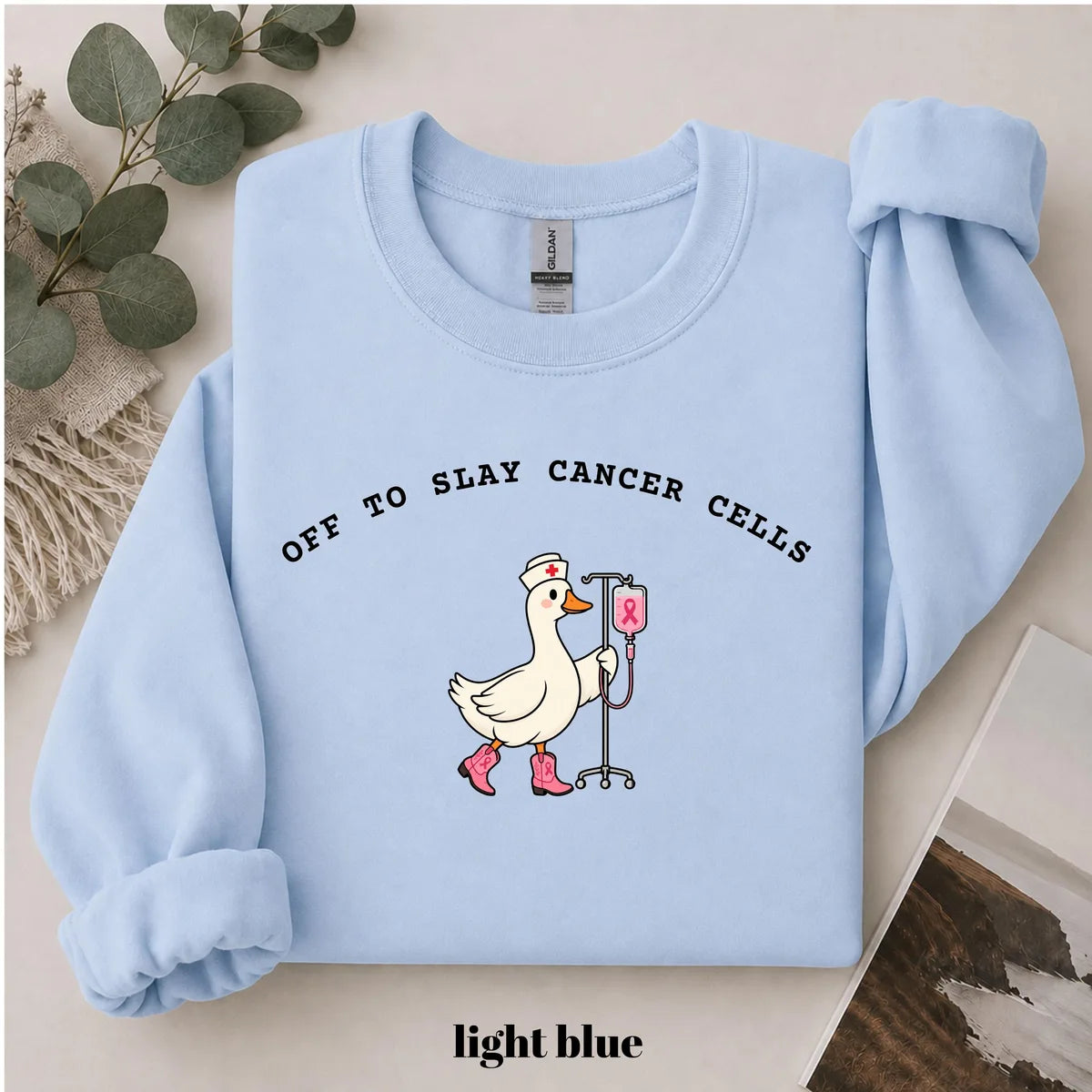 Light Blue crewneck sweatshirt with printed a nurse goose in pink boots with an IV pole and the words off to slay cancer cells, flat lay view.