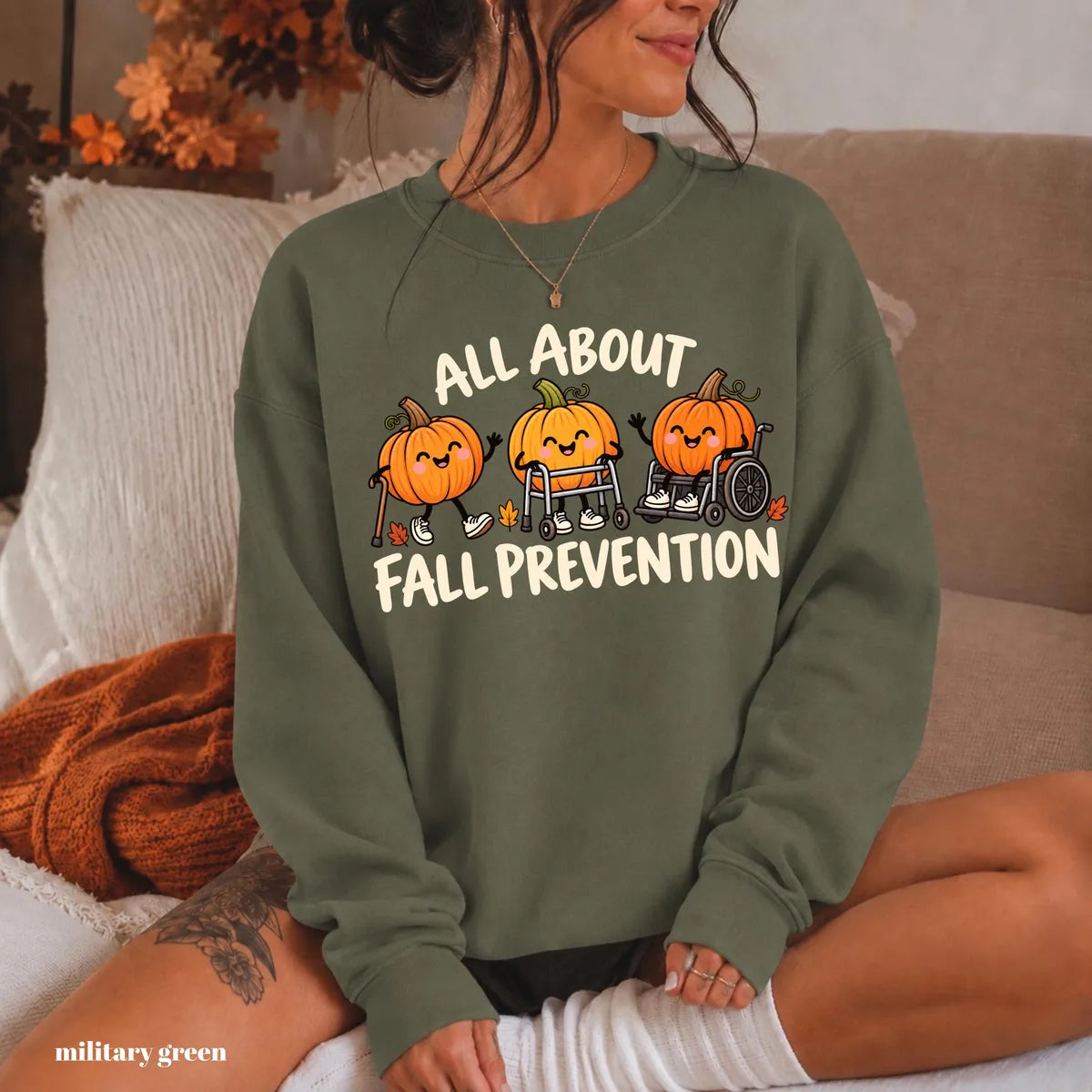 Military Green crewneck sweatshirt with printed pumpkins with a walker and wheelchair above the words all about fall prevention, worn by a model.