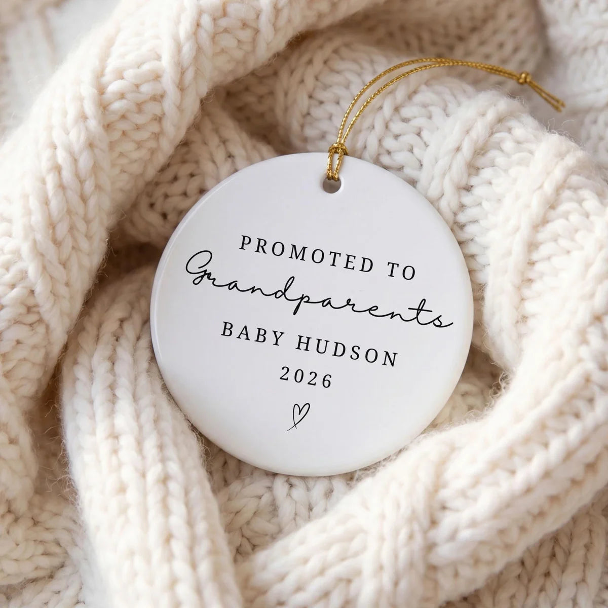 Round white ceramic Promoted to Grandparents ornament with a personalized baby name and year resting in a cream knit blanket