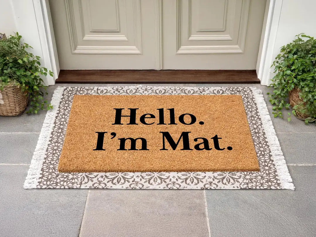 Coir doormat with a Hello I'm Mat message layered on a gray patterned fringed rug at a cream door