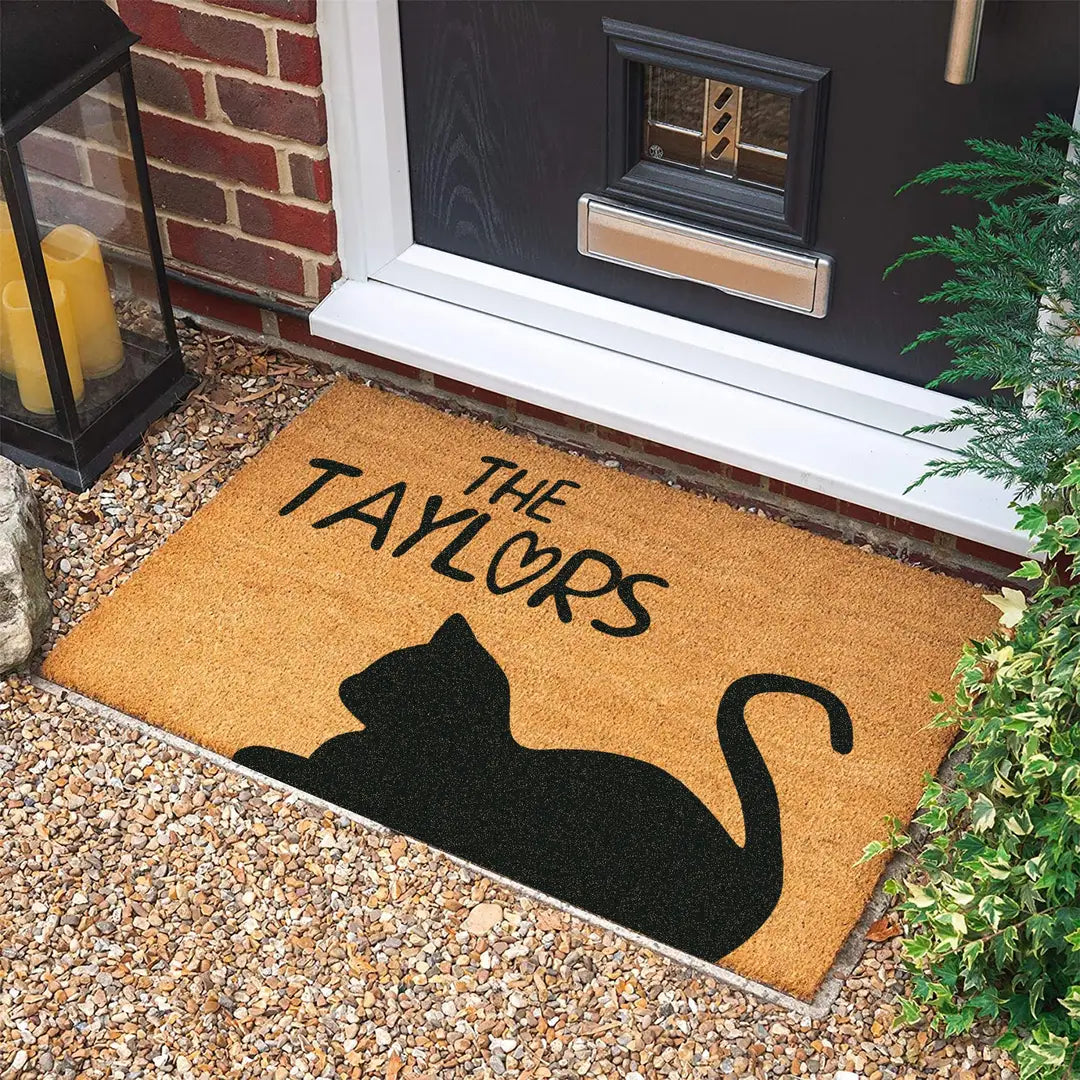 Coir doormat with a black cat silhouette and family name at a dark front door on gravel