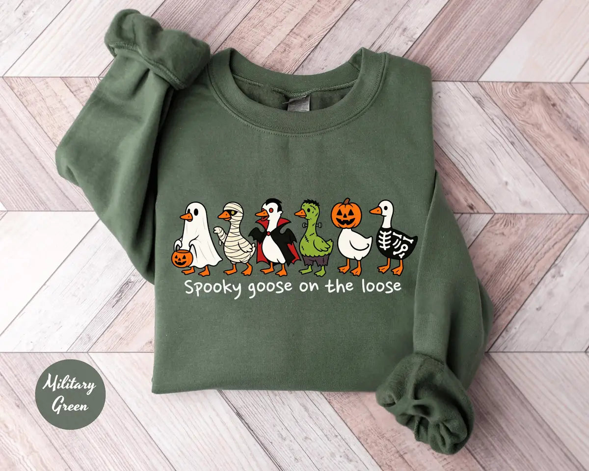 Military Green crewneck sweatshirt with costumed geese dressed as a ghost, vampire, pumpkin and skeleton above the words spooky goose on the loose, flat lay view.