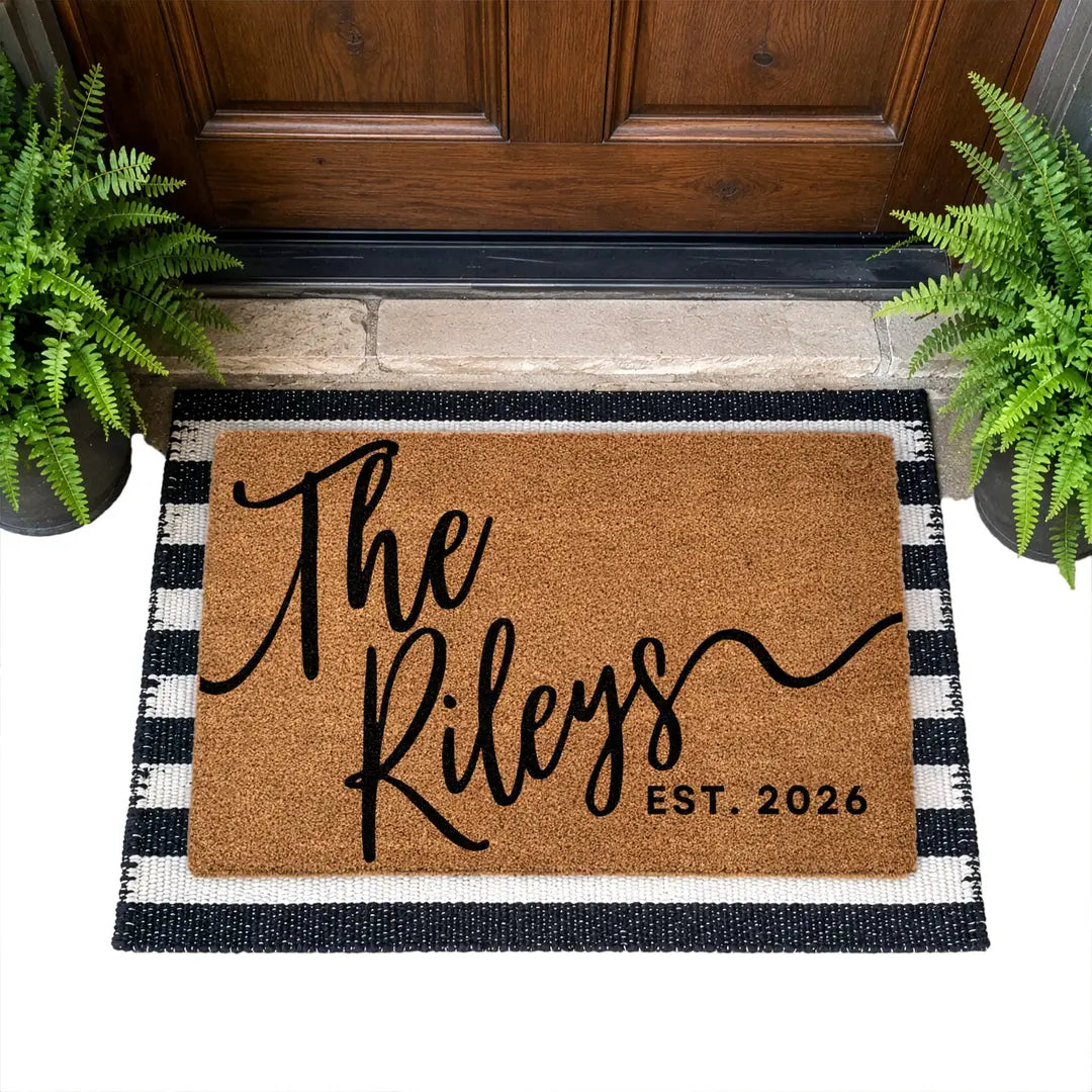 Coir doormat with a script family name and established year layered on a black and white striped rug at a wooden door with ferns