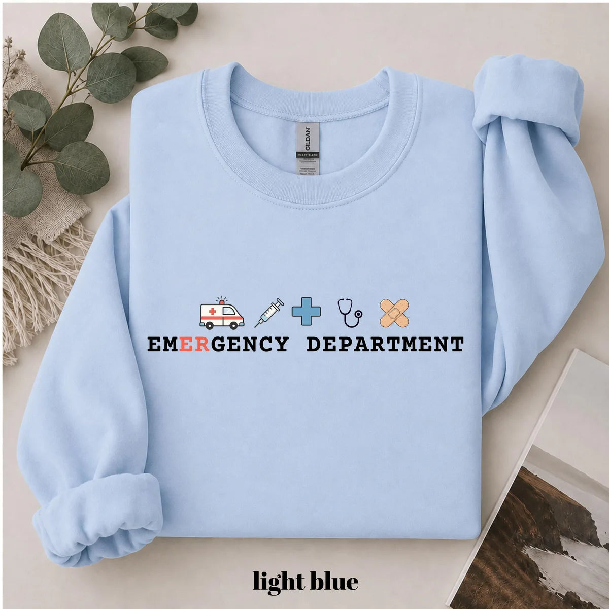 Light Blue crewneck sweatshirt with printed an ambulance, syringe, cross, stethoscope and bandage above the words emergency department, flat lay view.