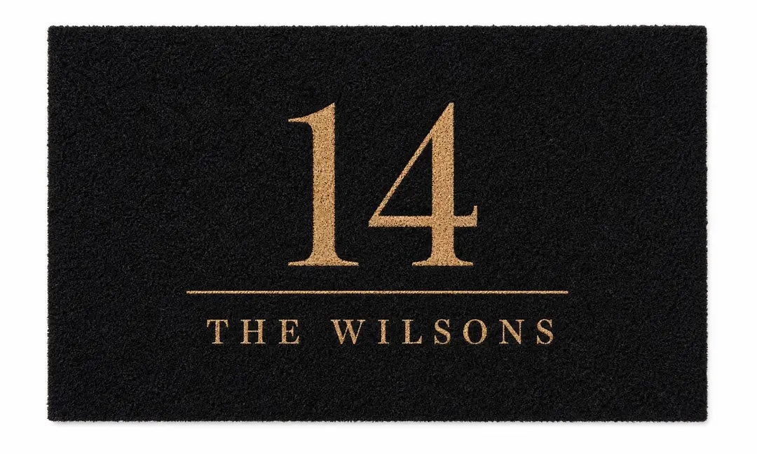 Flat view of a black faux coir doormat with a large gold-tone house number above a ruled family name