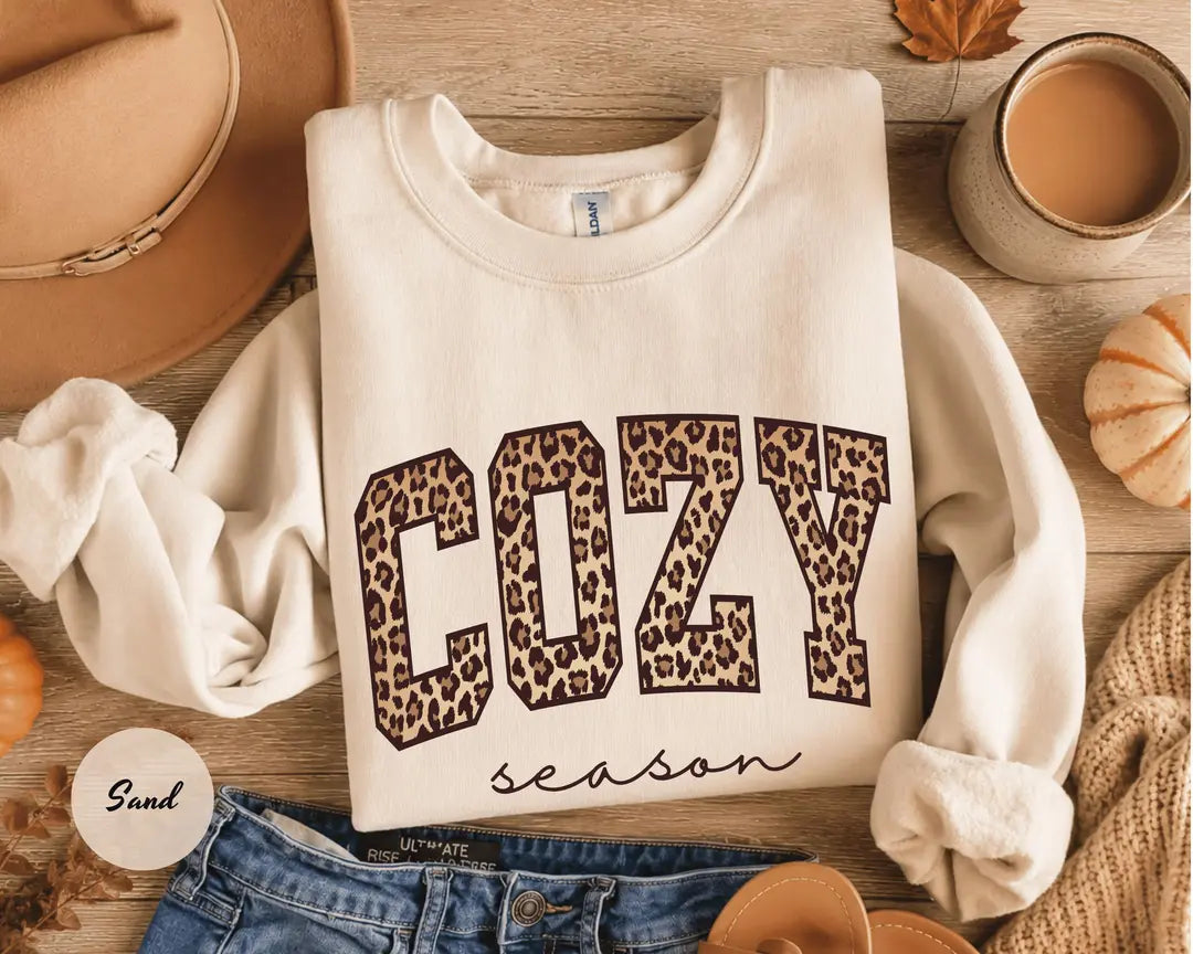 Sand crewneck sweatshirt with bold leopard print cozy lettering above the script words season, flat lay view.
