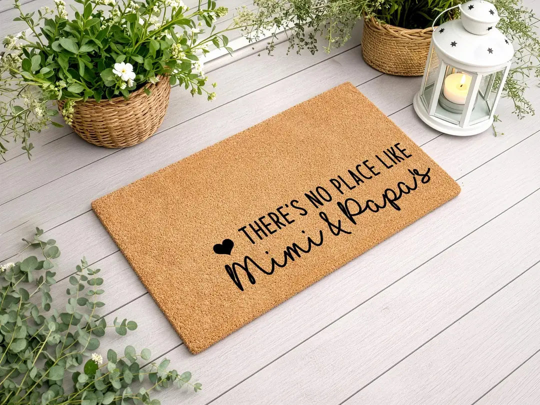 Coir doormat with a There's No Place Like Mimi and Papa's message on a white wood deck with a lantern and flower baskets