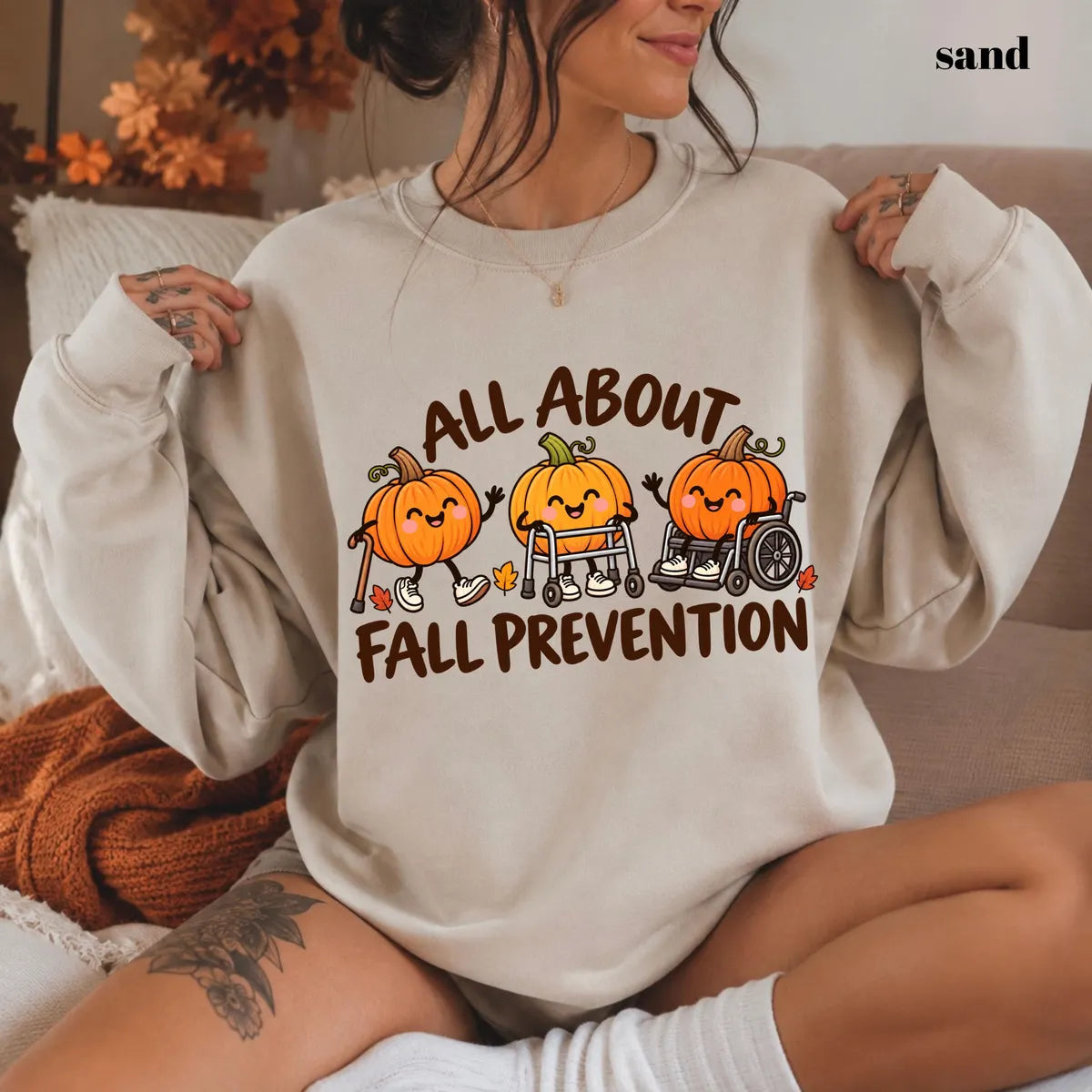 Sand crewneck sweatshirt with printed pumpkins with a walker and wheelchair above the words all about fall prevention, worn by a model.