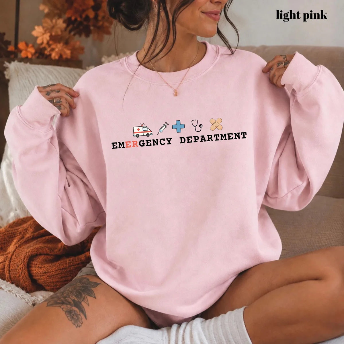 Light Pink crewneck sweatshirt with printed an ambulance, syringe, cross, stethoscope and bandage above the words emergency department, worn by a model.
